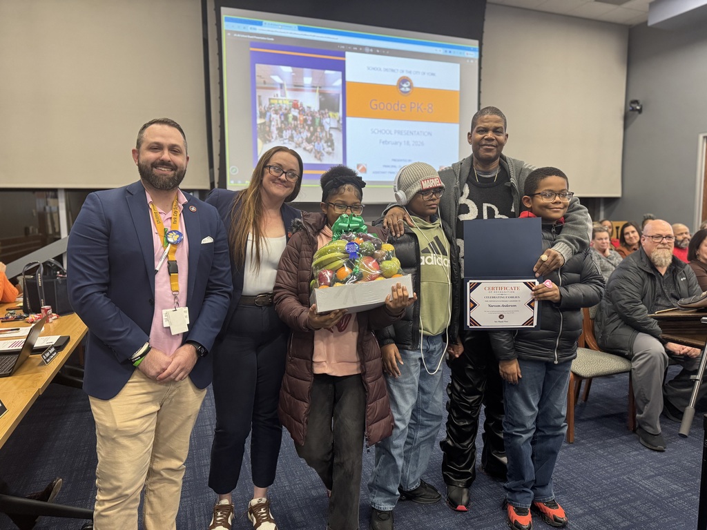 A group of six people, including Goode PreK-8 administrators and a family are standing in a conference room. A young girl is holding a fruit basket in her hand and the adult man is holding a certificate in his hand, with his arms wrapped around two young children.