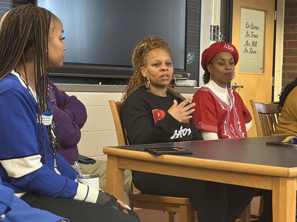 A district staff member along with various other adults are sitting around a table in a school library. The district staff member is speaking passionately.