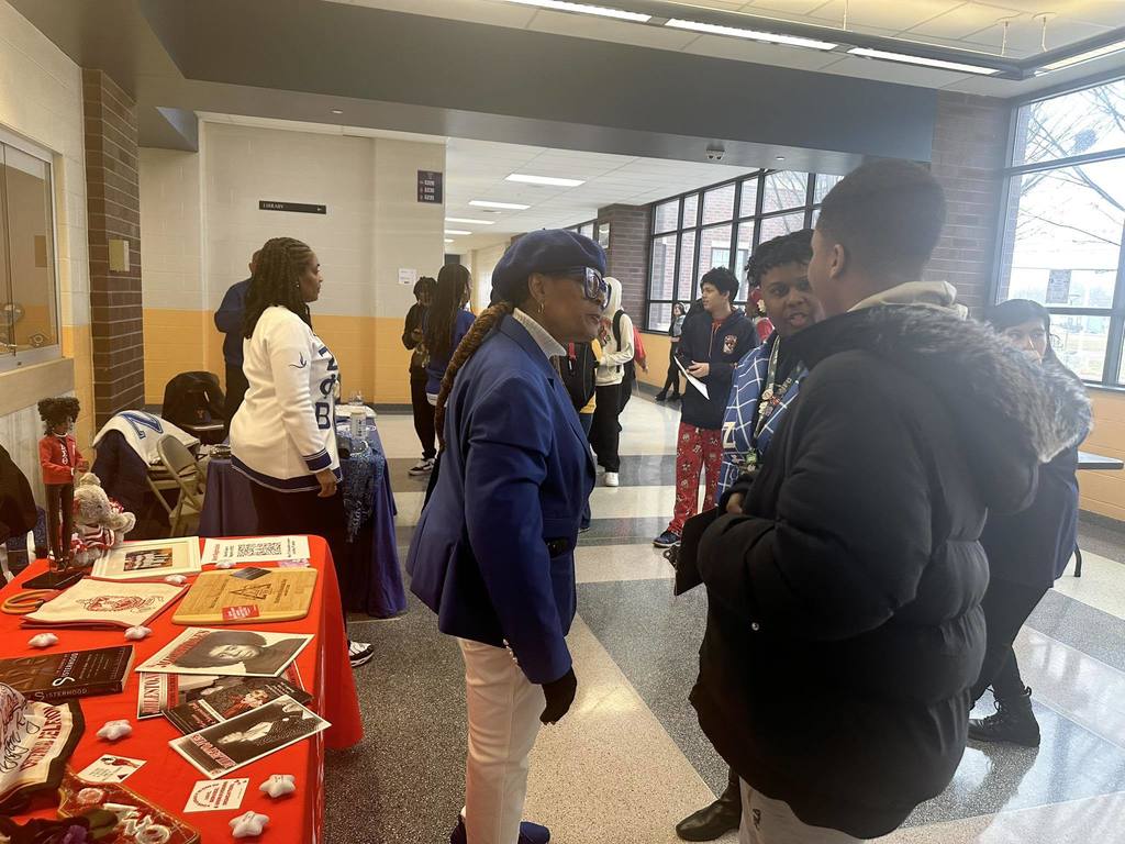 Two women are having a discussion with a student in a school hallway. Other people, including adults and students can be seen in the background.