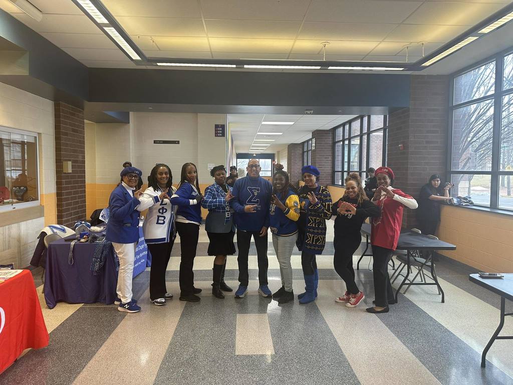 A group of nine adults are standing in a school hallway, holding up hand gestures. Tables and multiple students can be seen in the background.