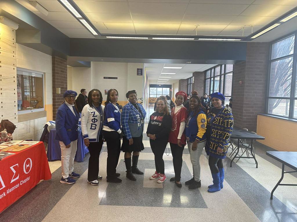 A group of eight adults are standing in a school hallway, holding up hand gestures. Tables and multiple students can be seen in the background.