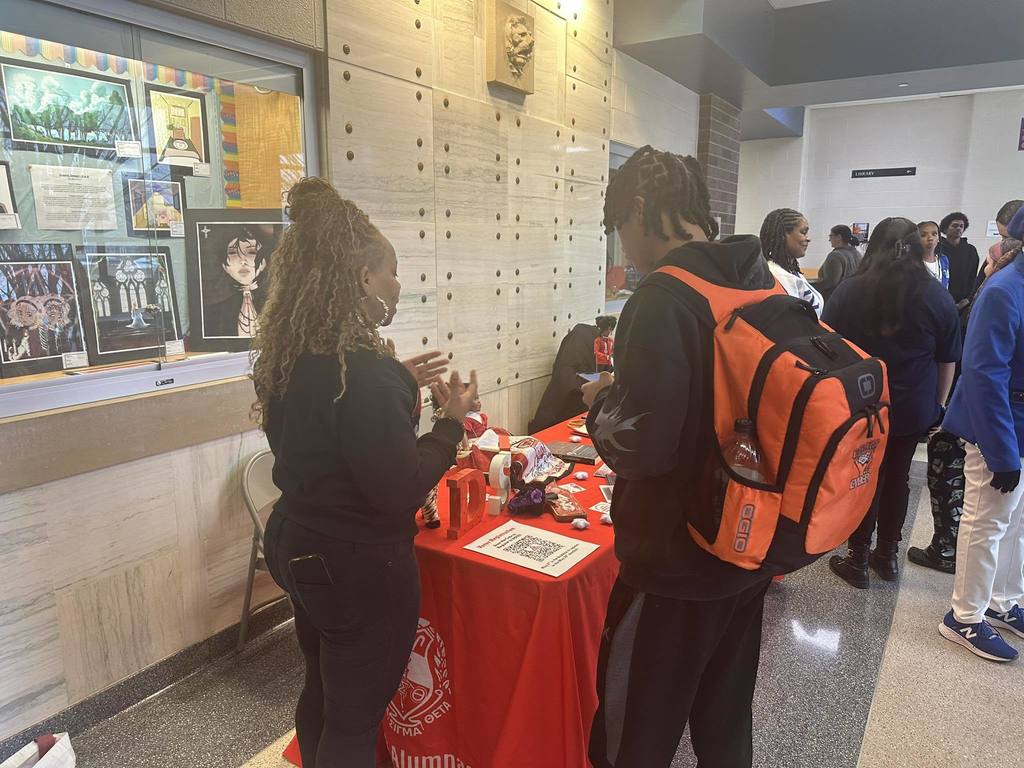 A district staff member is standing in a school hallway having a discussion with a student. Other students can be seen in the background.