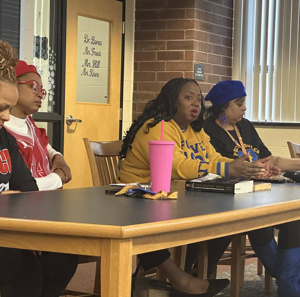 Four women are sitting at a table in a school library, engaging in a discussion. Books, snacks and a water bottle can be seen on the table.