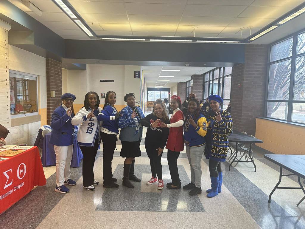 A group of eight adults are standing in a school hallway, holding up hand gestures. Tables and multiple students can be seen in the background.