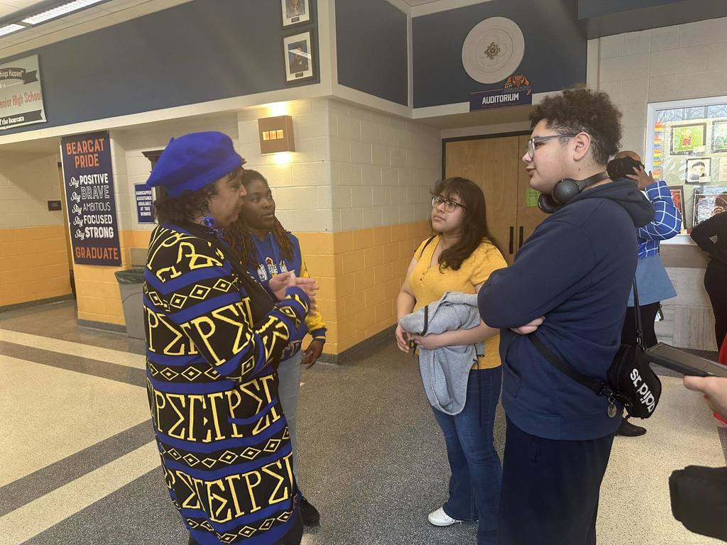 Two women are standing in a school hallway having a discussion with two students. Other students can be seen in the background.