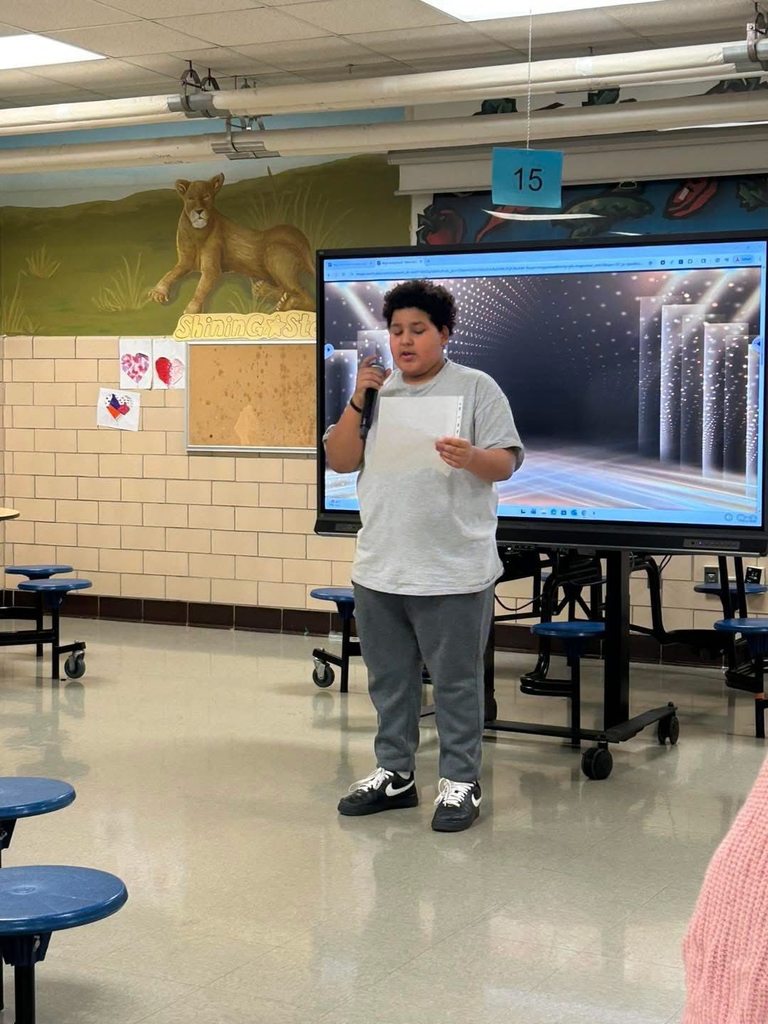 A young student is speaking into a microphone while holding a paper in his hand. A large screen displaying graphics can be seen nearby in the background.