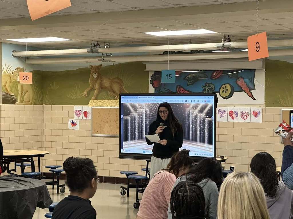 A student is speaking into a microphone while holding a paper in her hand. A large screen displaying graphics can be seen nearby in the background. Other people can be seen sitting at tables.