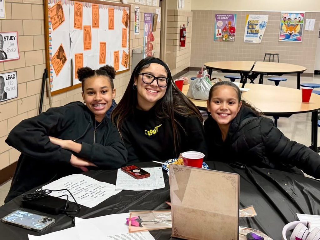Three students are sitting at a table in a school cafeteria. Papers, a phone, and a red cup can be seen on the table.