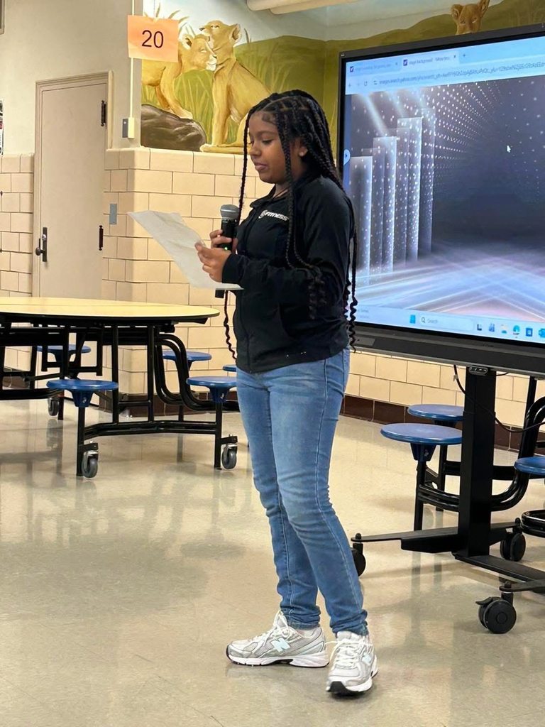 A young student is standing in a school cafeteria speaking into a microphone while holding a paper. A large screen displaying graphics can be seen nearby in the background.