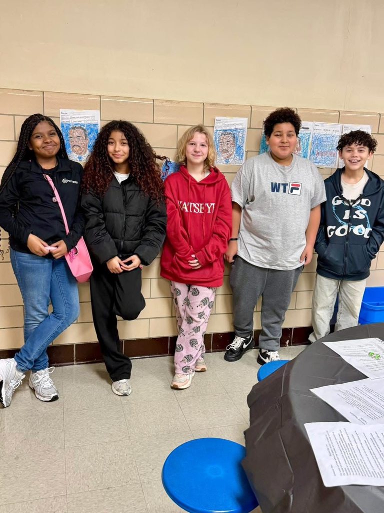 A group of five students are standing side by side in front of a wall showcasing artwork in a school cafeteria. 