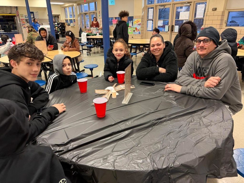 A family of five are sitting around a table in a school cafeteria. Red plastic cups are visible on the table. Other people can be seen in the background.