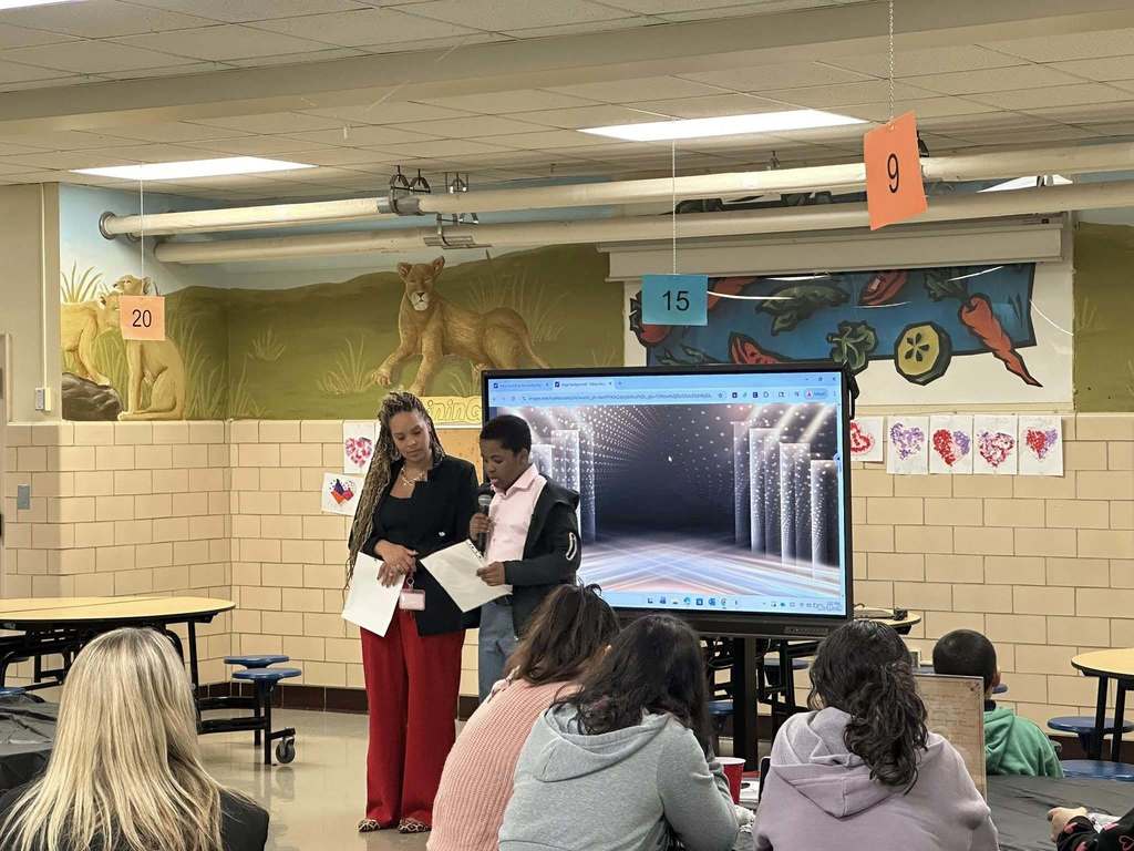 Devers PreK-8 principal, Dr. Hanson is standing next to a young student in a school cafeteria. The student is speaking into a microphone while holding a paper in his hand. A large screen displaying graphics can be seen nearby in the background. Other people can also be seen sitting at tables.