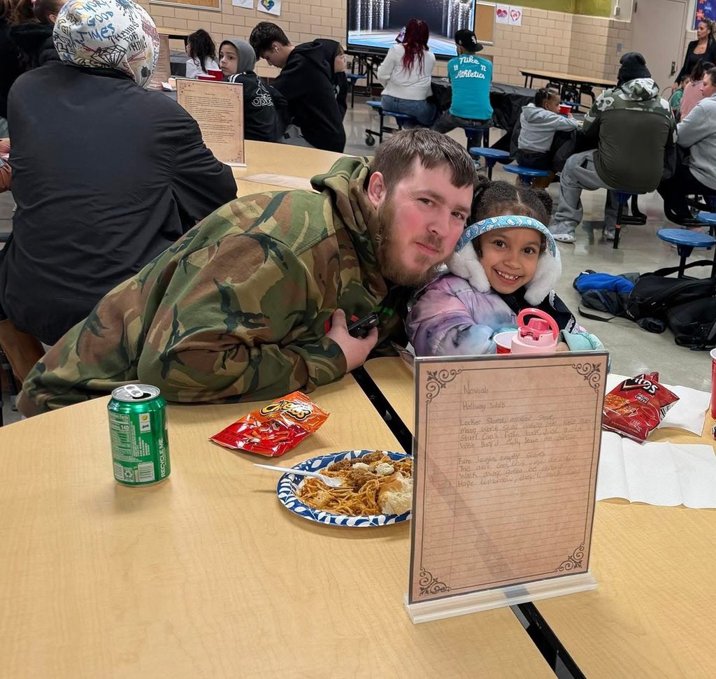 A man and a young girl are sitting at a table that has food and drinks on it in a school cafeteria. Other people can be seen in the background.