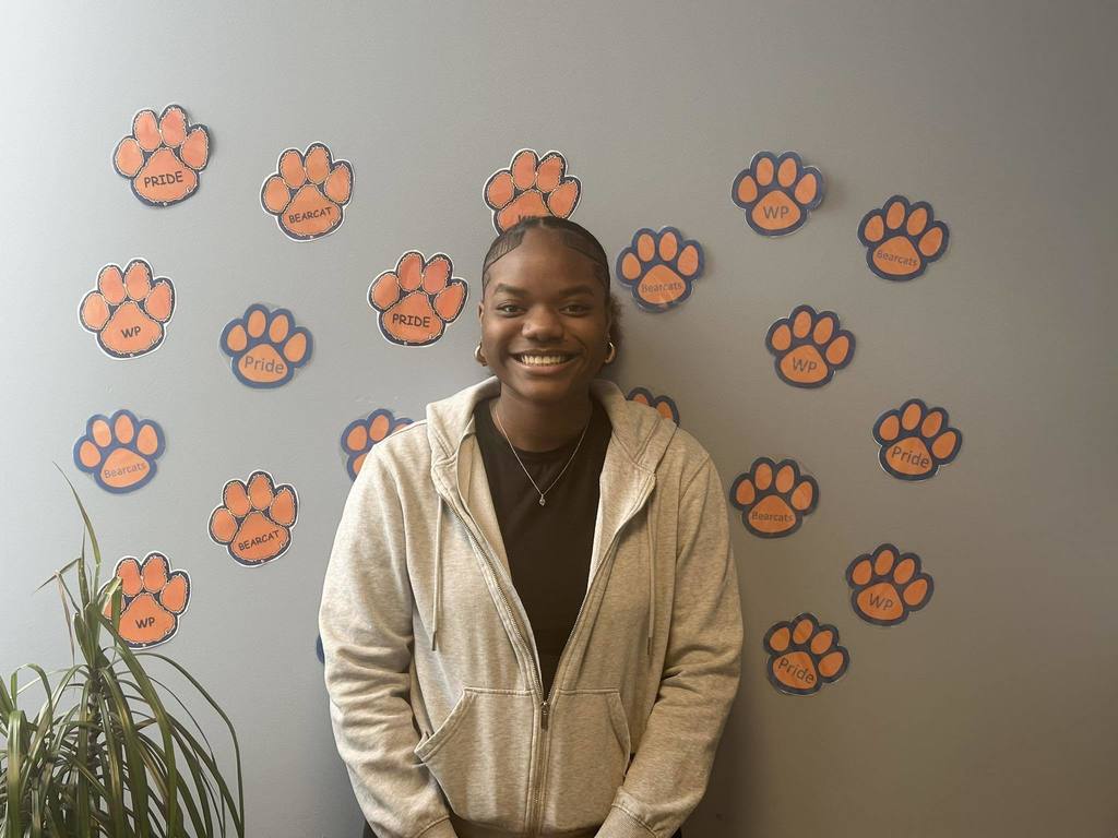 A student is standing in front of a wall decorated with orange paw prints labeled "Pride" and "Bearcat." A plant is visible on the left.