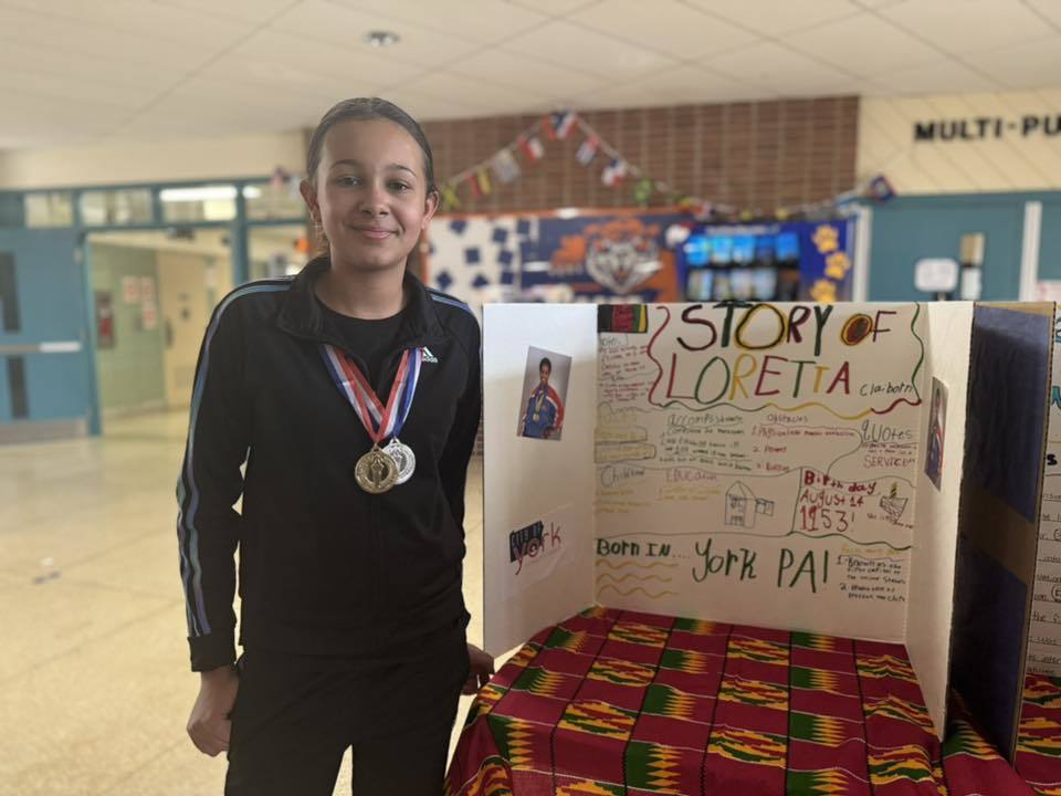 A student is standing in a school hallway, next to a tri-fold display titled "Story of Loretta Claiborne," featuring text, illustrations, and photos on a patterned tablecloth.