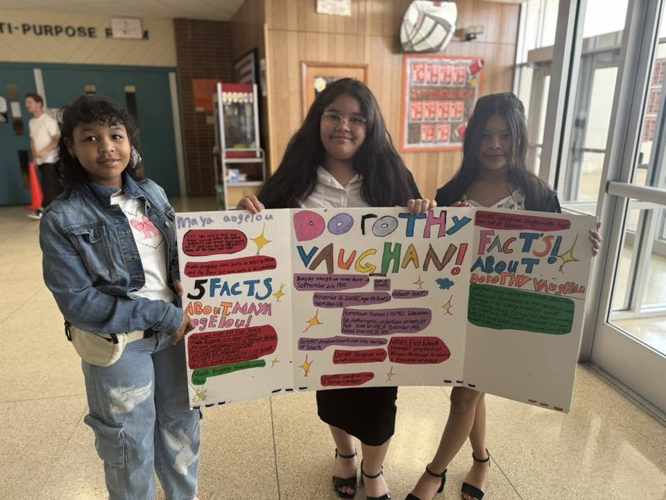 A group of three students are standing in a school hallway, holding a colorful poster about Dorothy Vaughn.