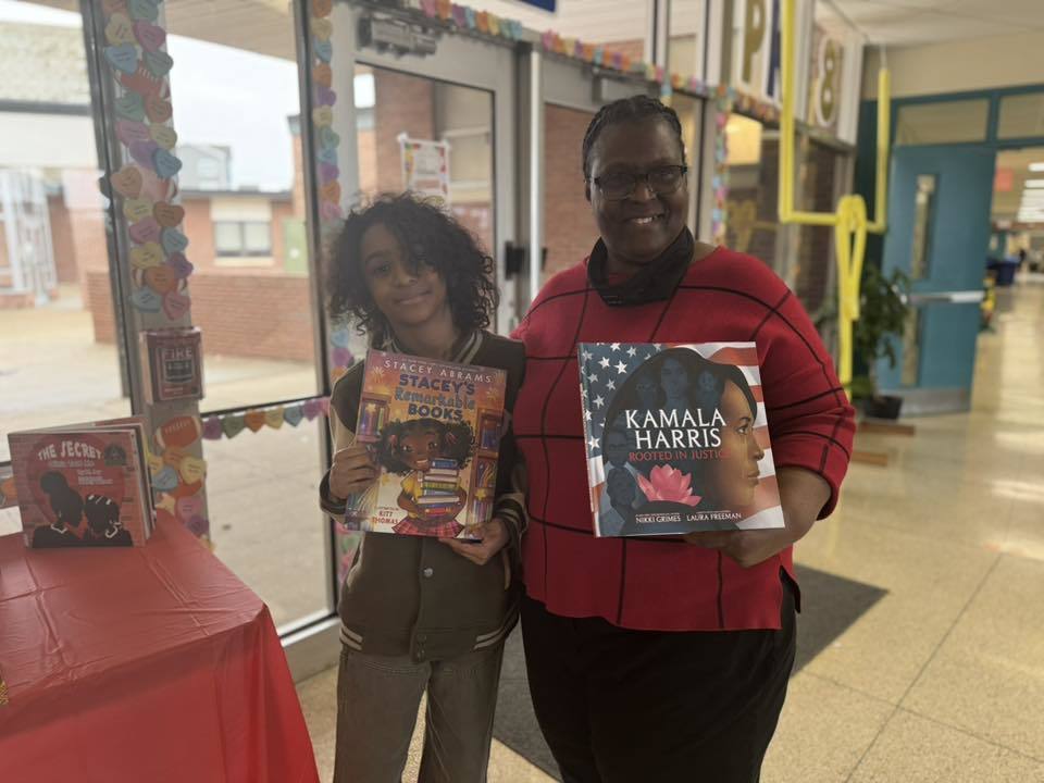 A district staff member and a student are standing in a school hallway holding books titled "Stacey Abrams: Stacey's Extraordinary Words" and "Kamala Harris: Rooted in Justice."
