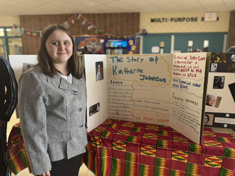 A student is standing in a school hallway, next to a tri-fold display titled "The Story of Katherine Johnson," featuring text, illustrations, and photos on a patterned tablecloth.