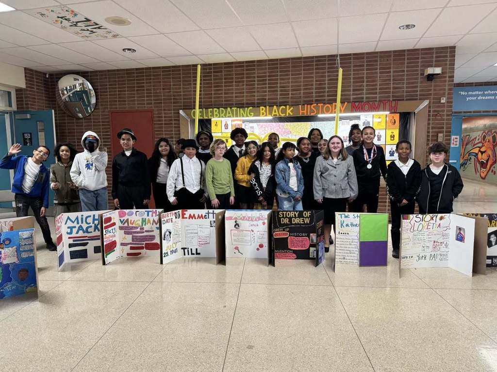 A group of students are standing in a school hallway celebrating Black History Month. They are displaying colorful projects on notable figures.