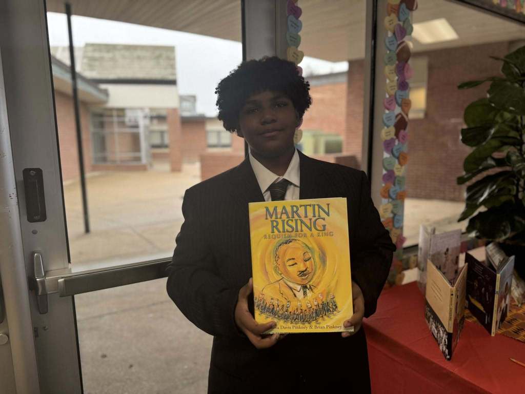 A student is standing in a school hallway, holding a book titled "Martin Rising." Books and a heart-decorated window can be seen in the background.