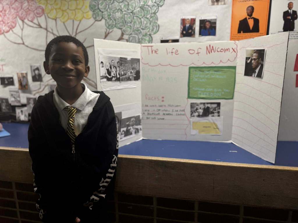 A student is standing in a school hallway, next to a tri-fold display titled "The life of Malcolm X," featuring text, illustrations, and photos.