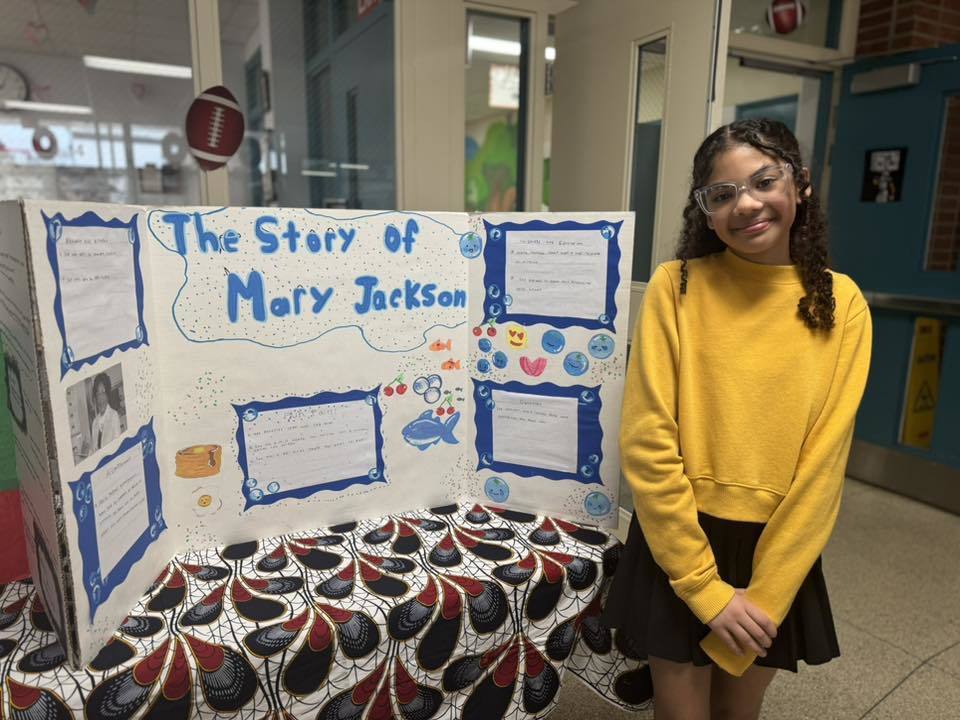 A student is standing in a school hallway, next to a tri-fold display titled "The Story of Mary Jackson," featuring text, illustrations, and photos on a patterned tablecloth.
