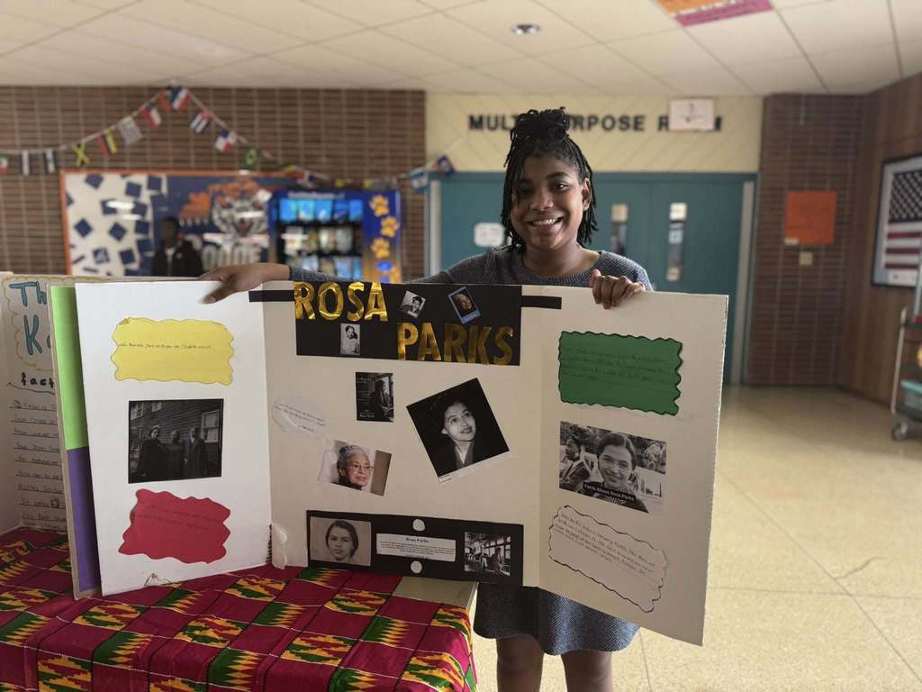 A student is holding a tri-fold poster about Rosa Parks, featuring photos and text, while standing in a school hallway decorated with colorful banners.