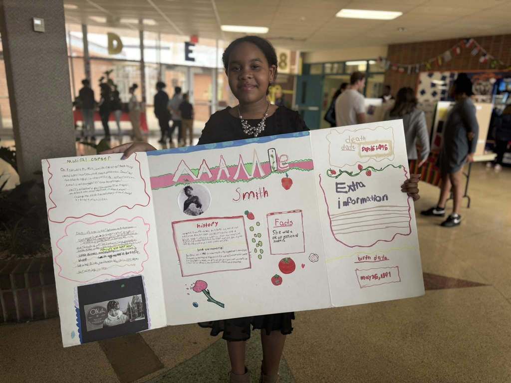A student is standing in a school hallway, holding a tri-fold poster with hand-drawn graphics and text. Other people and displays can be seen in the background.