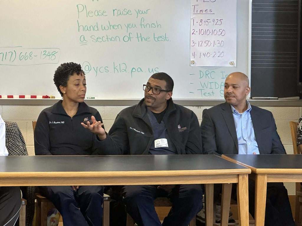 A diverse panel of community members sitting at a table engaging in discussion in a school library. 