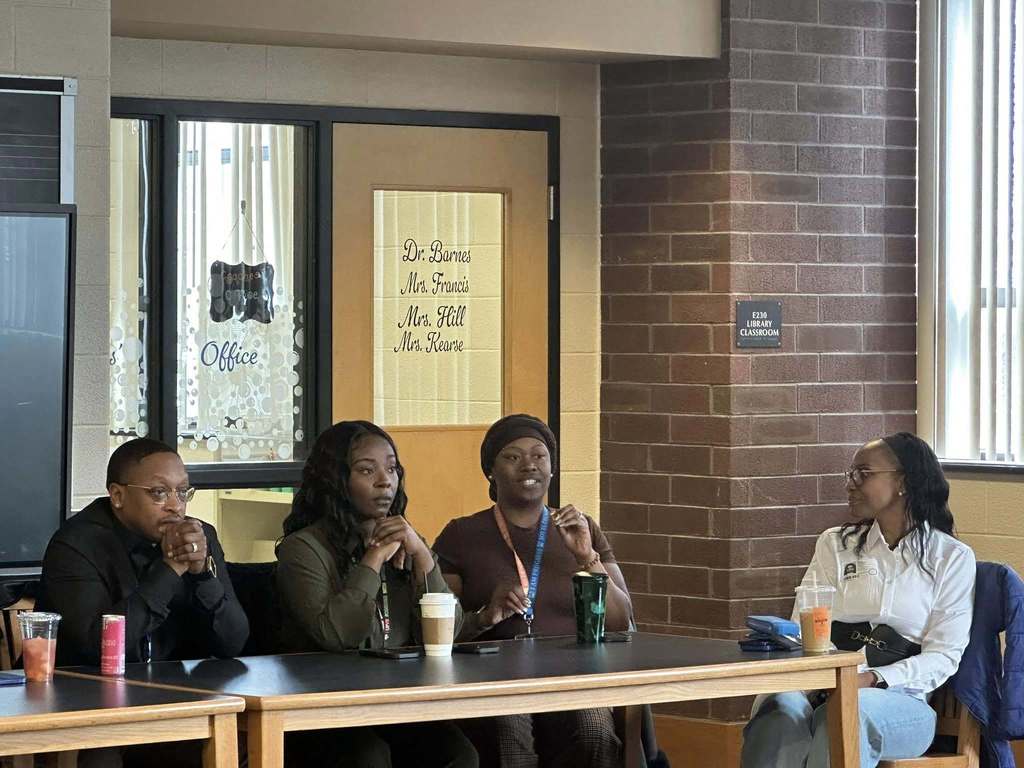 A diverse panel of community members sitting at a table engaging in discussion in a school library. 