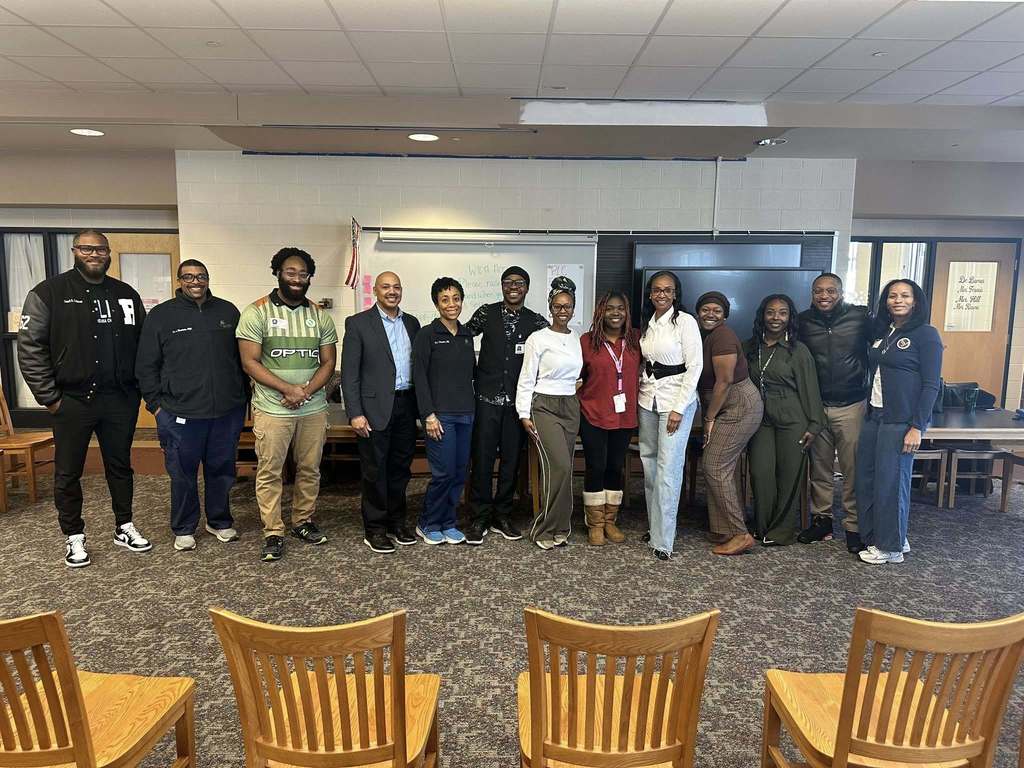 A group of thirteen adults, including various community members and a district staff member, are standing in a school library surrounded by a whiteboard and chairs. 