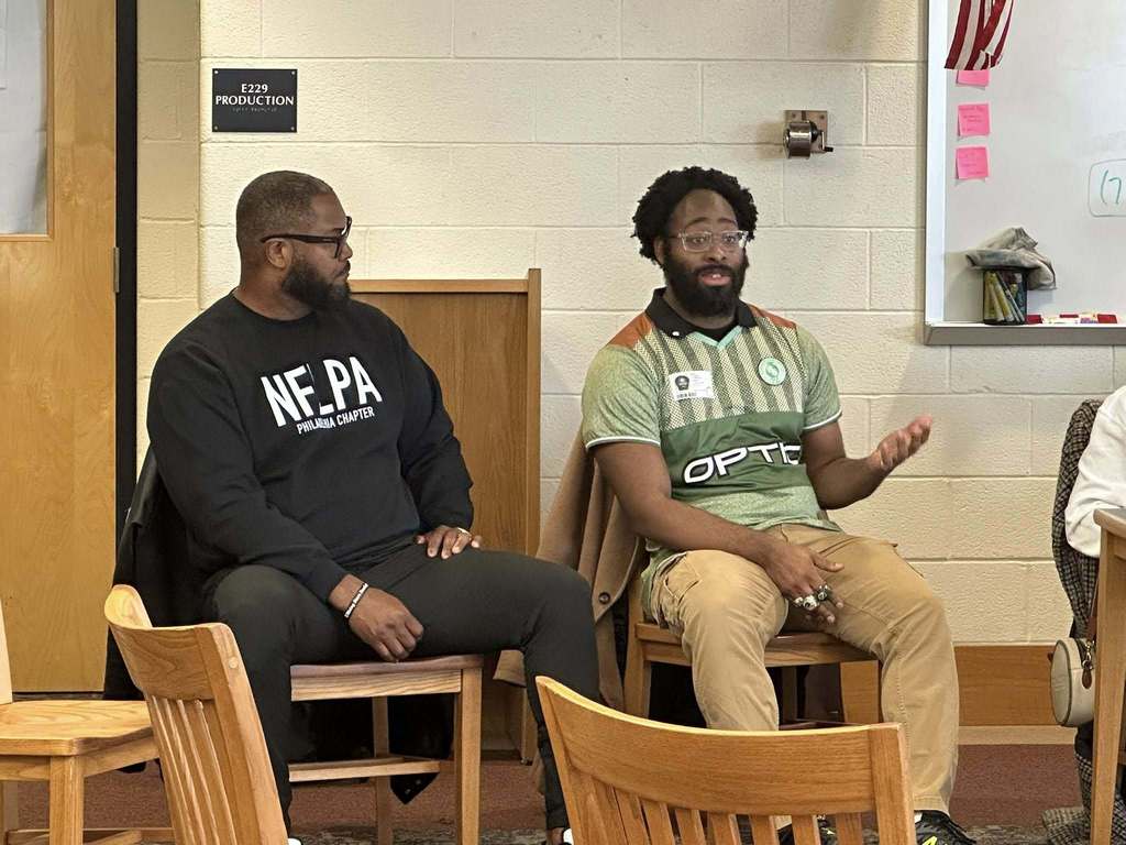 A diverse panel of community members sitting at a table engaging in discussion in a school library. 