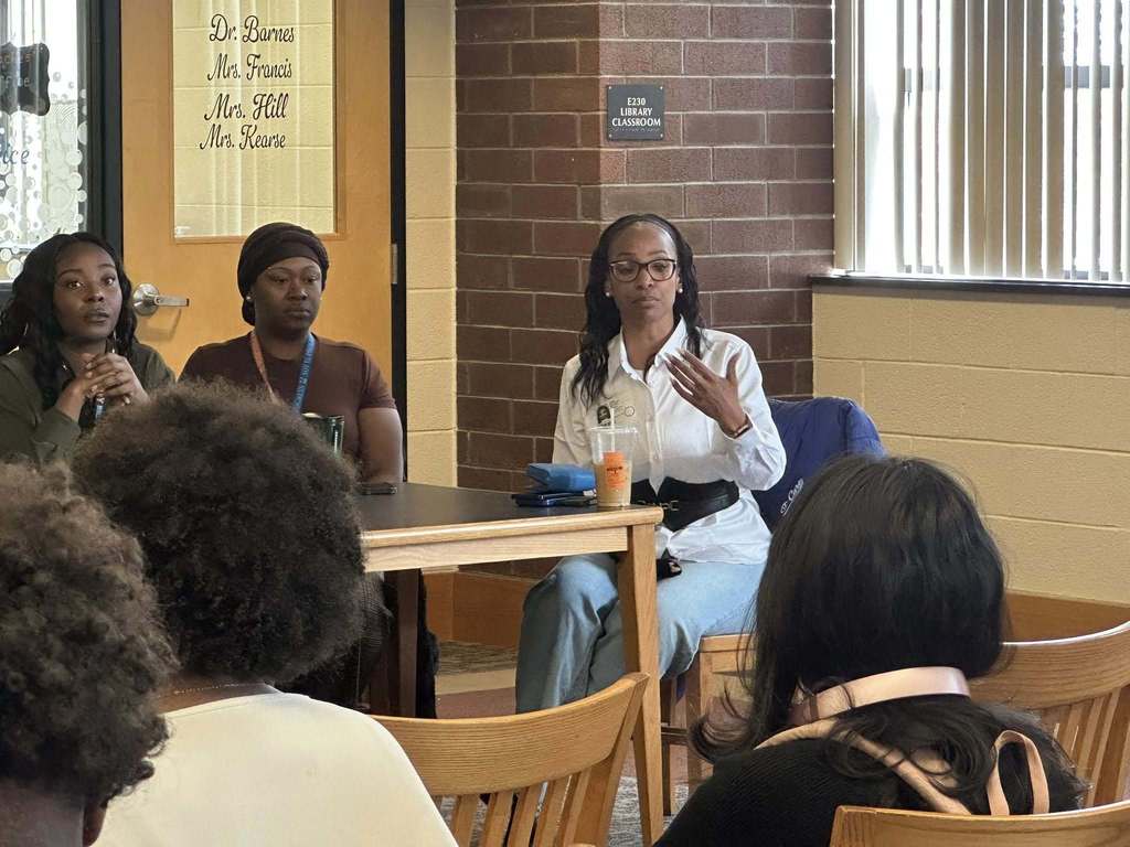 A diverse panel of community members sitting at a table engaging in discussion in a school library. 