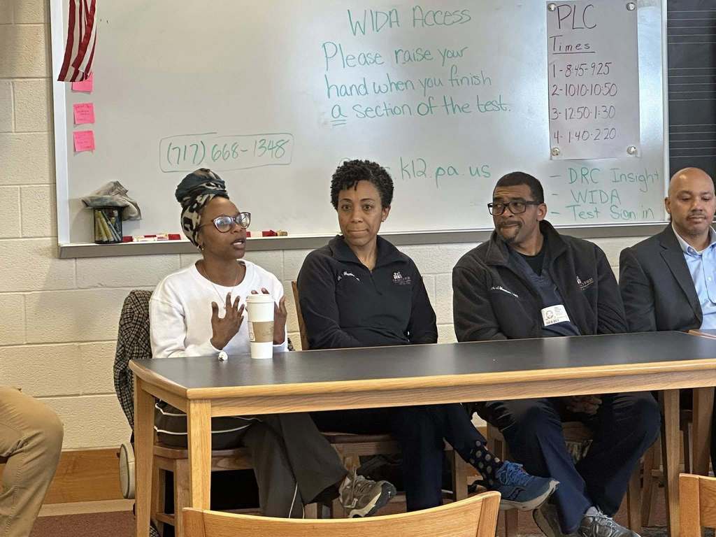 A diverse panel of community members sitting at a table engaging in discussion in a school library. 