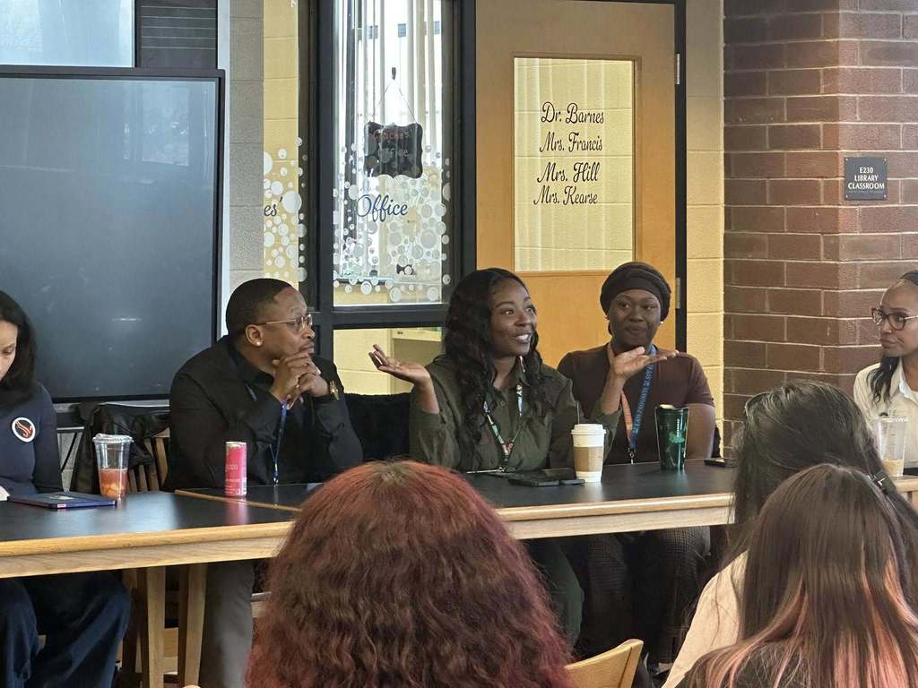 A diverse panel of community members sitting at a table engaging in discussion in a school library. 