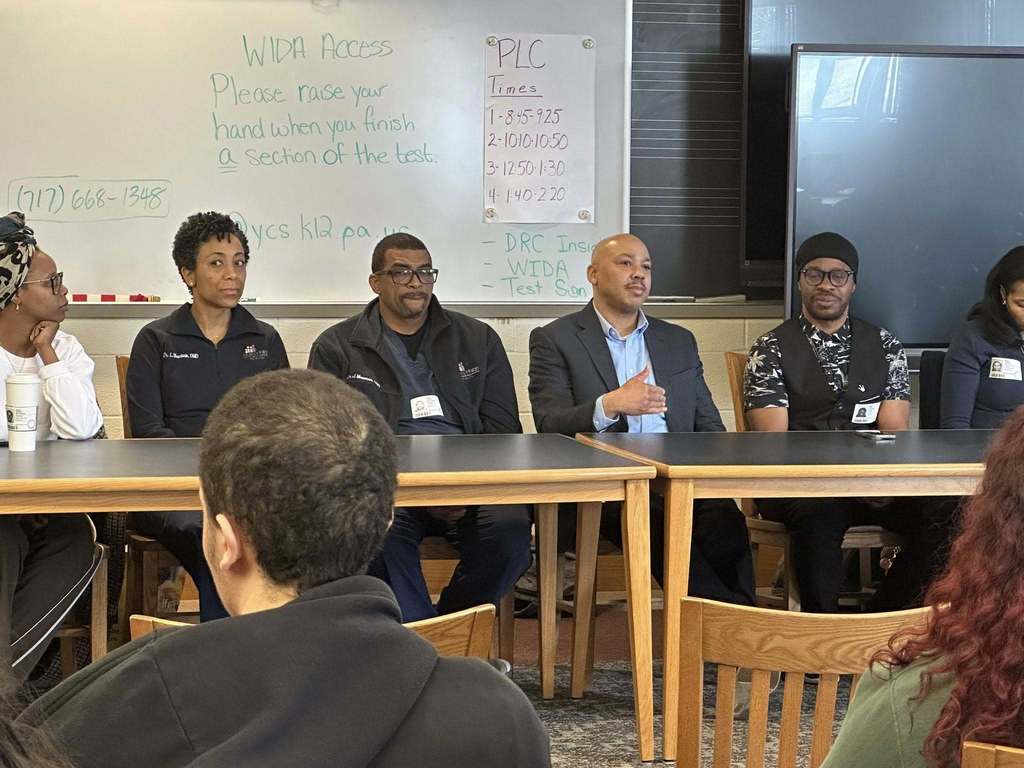 A diverse panel of community members sitting at a table engaging in discussion in a school library. 
