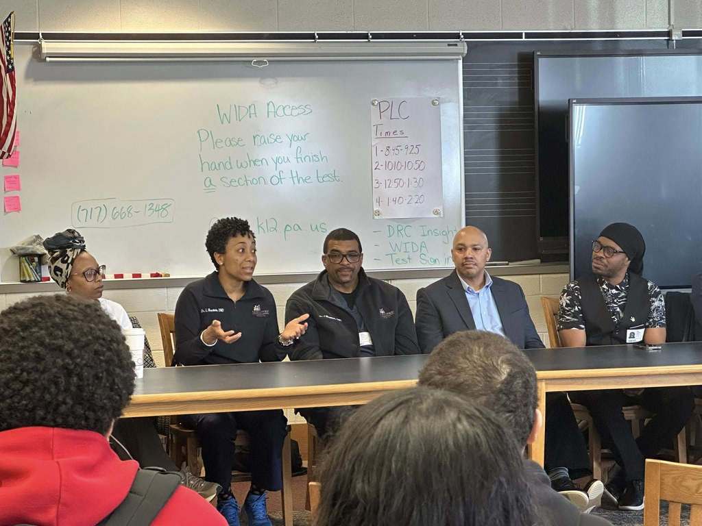 A diverse panel of community members sitting at a table engaging in discussion in a school library. 