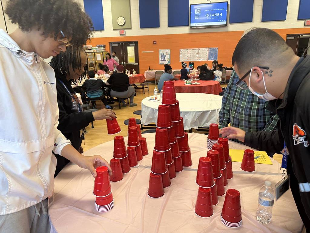 A group of three people, including two district staff members, are standing around a table in a school cafeteria, building a pyramid with red plastic cups. Other people can be seen sitting at tables nearby in the background.