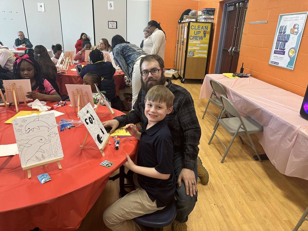 An adult man is kneeling down next to a young boy who is sitting at a table with art supplies on it in a school cafeteria. Other people can be seen nearby in the background.