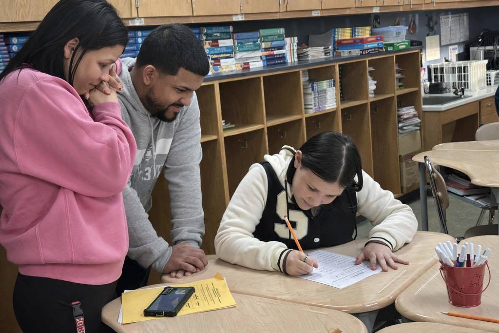A woman and a man are looking down at a girl who is sitting at a table in a school classroom. The girl is writing on a piece of paper with a pencil. Shelves of books can be seen nearby in the background.