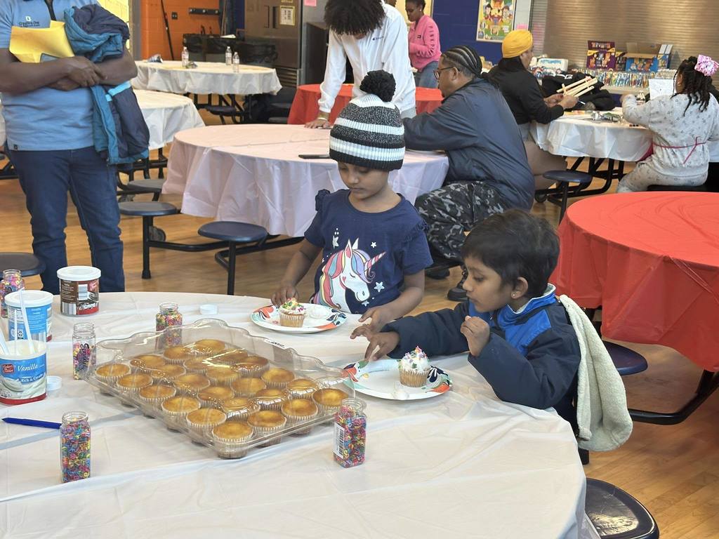 Two young children are sitting at a table in a school cafeteria decorating cupcakes with sprinkles and frosting. Other people can be seen nearby in the background.