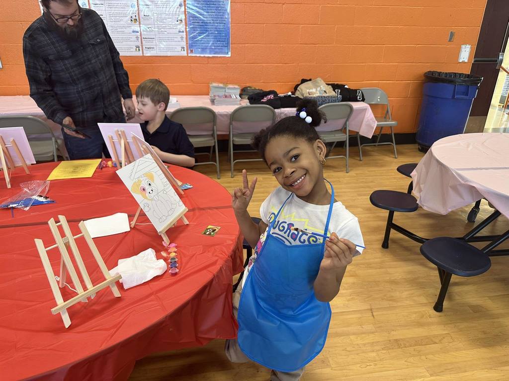 A young girl in a blue apron is holding up a peace sign while standing beside a table with art supplies in a school cafeteria. A man and a young boy can be seen sitting nearby in the background.