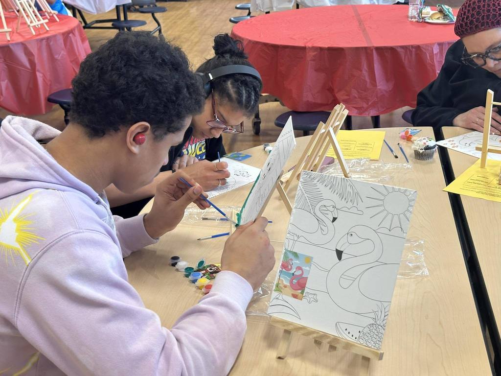 Two teenagers and an adult woman are painting canvases while sitting at a table in a school cafeteria. Paint pots and brushes are scattered on the table.