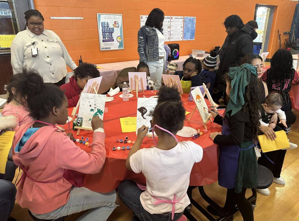 A group of children are sitting around a table in a school cafeteria, engaging in painting canvases. Adults can be seen nearby in the background.