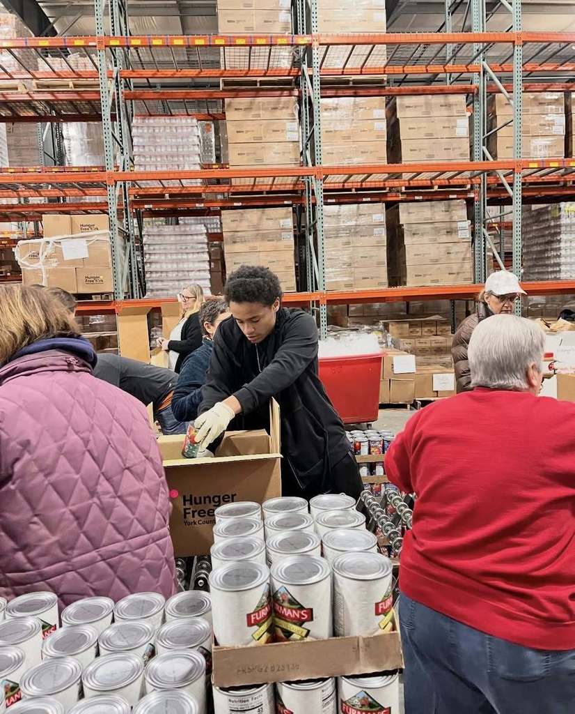 A group of volunteers, including a student volunteer, are packing canned goods in a warehouse that has tall shelves of boxes.