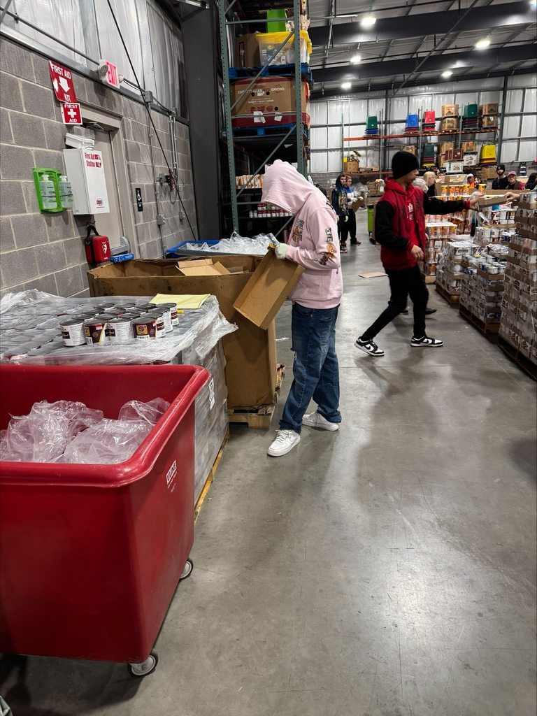 A student volunteer is inspecting boxes in a warehouse with canned goods. Other people can be seen nearby in the background.