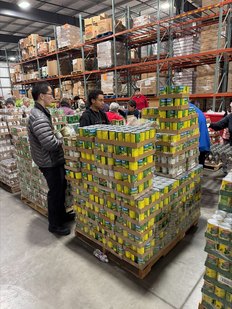 A group of student volunteers are standing in a warehouse, stacking canned goods on pallets. Shelves with boxed supplies can be seen in the background.