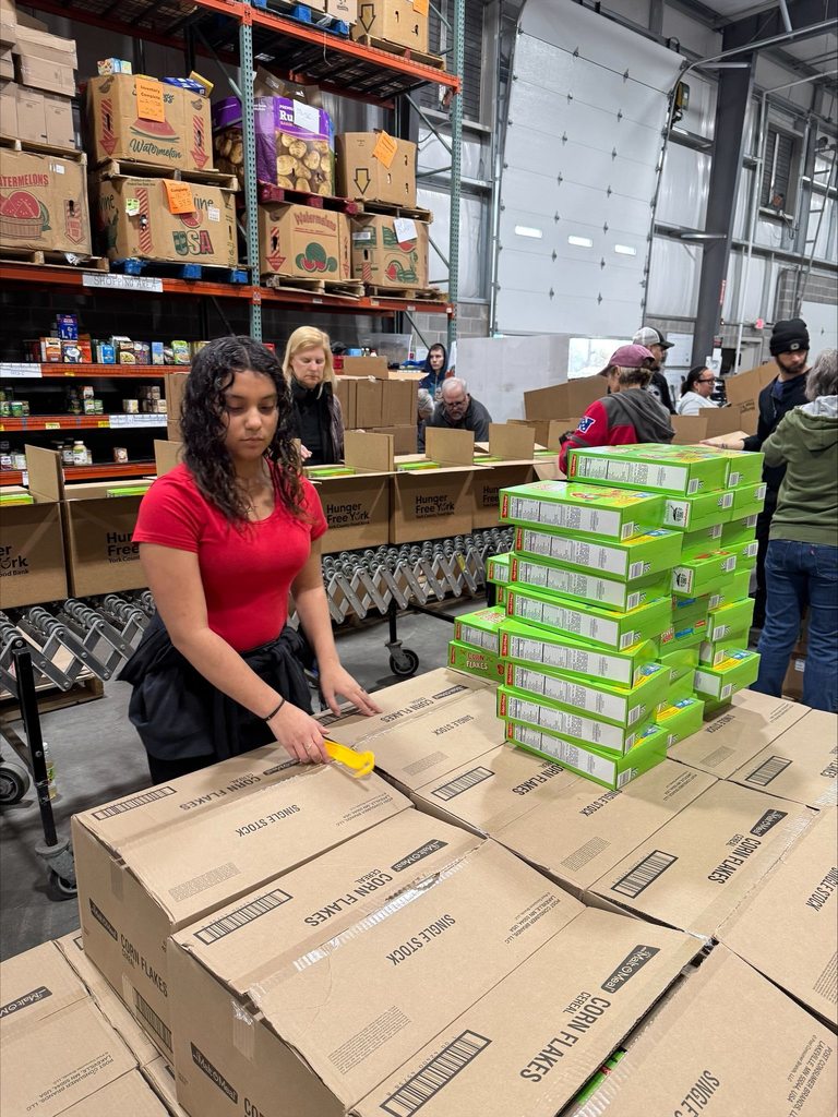 A student volunteer is packing boxes in a warehouse filled with stacks of food supplies. Other people can be seen nearby in the background.
