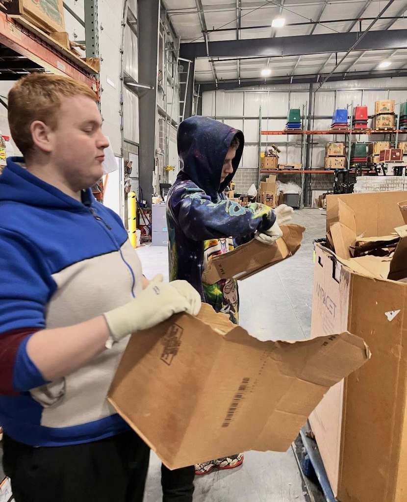 Two student volunteers are standing in a warehouse sorting through cardboard boxes. Shelves with boxes can be seen in the background.