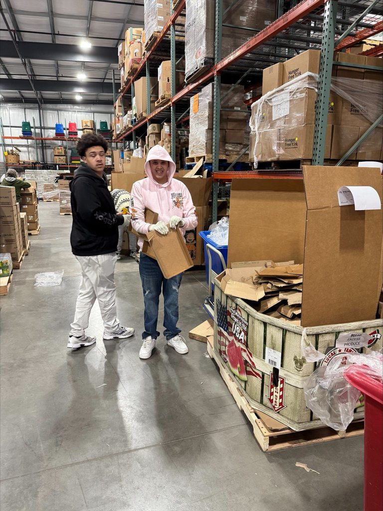 Two student volunteers are standing in a warehouse organizing cardboard boxes. The shelves are stocked with various items. 